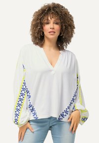 Hvid langærmet blouse med V-hals, der har blå og gule geometriske mønstre langs ærmerne og en knapdetalje ved halsudskæringen.