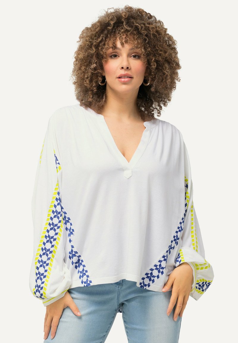 Hvid langærmet blouse med V-hals, der har blå og gule geometriske mønstre langs ærmerne og en knapdetalje ved halsudskæringen.