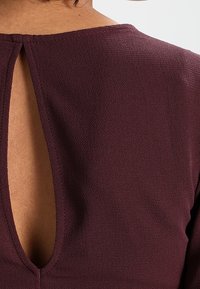 Liliowa bluzka o teksturowym wykończeniu z dekoltem w kształcie dziurki. Dekolt z przodu jest wysoki, z małym otworem z tyłu.