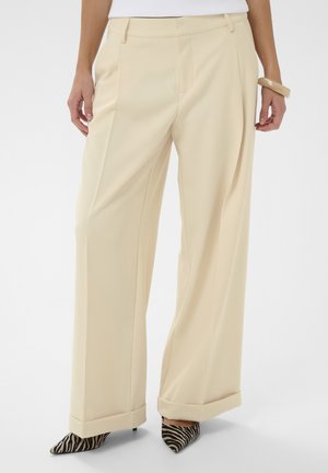 Kobieta ubrana w beżowe spodnie z wysokim stanem i szerokimi nogawkami, zestawione z butami na obcasie w paski zebry oraz beżową bransoletką na prawej ręce.