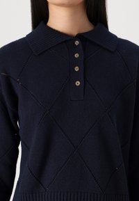 Dunkelblauer Strickpullover mit Rautenmuster, geripptem Kragen, vierknöpfiger Knopfleiste und geripptem Saum, getragen von einer Person mit langen dunklen Haaren.