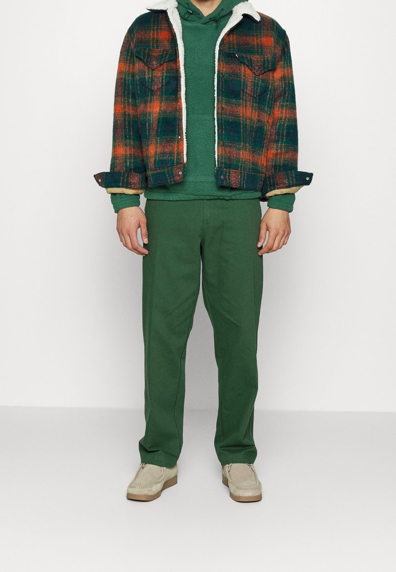 Homme portant un pantalon vert, un sweat à capuche vert et une veste à carreaux verte et orange avec des chaussures beiges, debout devant un fond blanc.