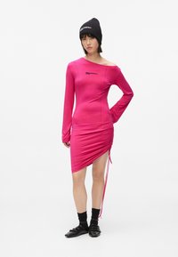 Γυναίκα που φοράει ένα εφαρμοστό φανταχτερό ροζ φόρεμα με πτυχές, μαύρο σκουφάκι, μαύρες κάλτσες και μαύρα παπούτσια με αγκώνα, στέκεται με το ένα χέρι στη μέση.