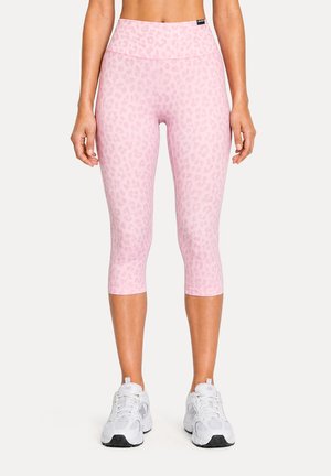 Femme portant des leggings courts à motif léopard rose et des chaussures de sport blanches, debout devant un fond uni.