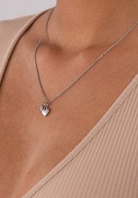 Silberne Halskette mit einem kleinen Herzanhänger und einem dekorativen Akzent, getragen vor einem neutralen Stoffhintergrund.