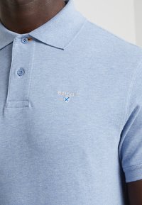 Světle modré polo tričko z texturované látky, se stojáčkem, dvěma knoflíky a vyšitým logem 'Barbour' na hrudi.