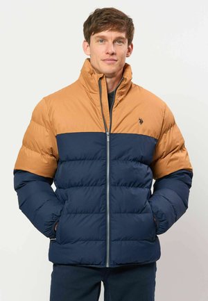 Man wearing a two-tone padded jacket with tan upper and navy lower sections, standing with hands in pockets against a plain background.