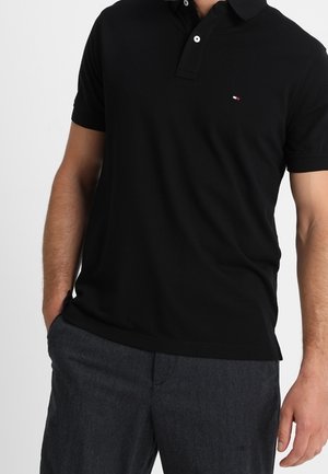 Homme portant un polo à manches courtes noir et un pantalon gris foncé, une main dans la poche et avec un petit logo sur la poitrine.