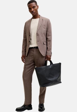 Man in een bruin pak en een beige overhemd, die een grote zwarte leren totebag vasthoudt, staand tegen een effen lichte achtergrond.