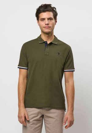 Mann mit kurzen dunklen Haaren, trägt ein olivgrünes Poloshirt mit gestreiften Ärmelbündchen und beigefarbene Hose, steht vor einem einfarbigen Hintergrund.