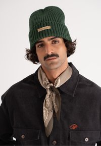 Grøn ribbet hue med et logo-tag; båret med en mørk corduroy skjorte med en brystlomme og et beige mønstret tørklæde bundet om halsen.