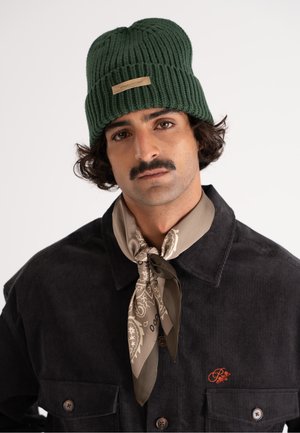 Green ribbed beanie with a logo tag; worn with a dark corduroy shirt featuring a chest pocket and a beige patterned scarf tied around the neck.