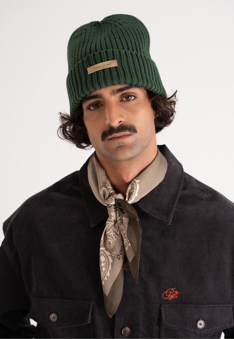 Beanie verde cu nervuri și etichetă cu logo; purtat cu o cămașă din catifea întunecată, care are un buzunar pe piept, și o eșarfă bej cu model legată în jurul gâtului.