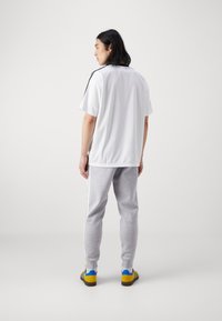 Bela atletična majica s kratkimi rokavi in črnimi poudarki, kombinirana s sivimi trenirkami s stranskimi črtami in rumenimi športnimi copati z modrimi poudarki.