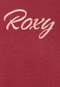 Roxy-logotypen broderad med ljusrosa tråd på röd tyg, som visar detaljerad struktur av sömmen och materialet.
