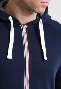 Sudadera con capucha azul marino con cordones blancos, que cuenta con un cierre frontal y detalles a rayas rojas y blancas a lo largo de la cremallera.