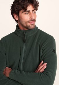 Dark green fleece jacket with a high collar, front zipper, and logo patch on the sleeve. Soft texture, fitted design.