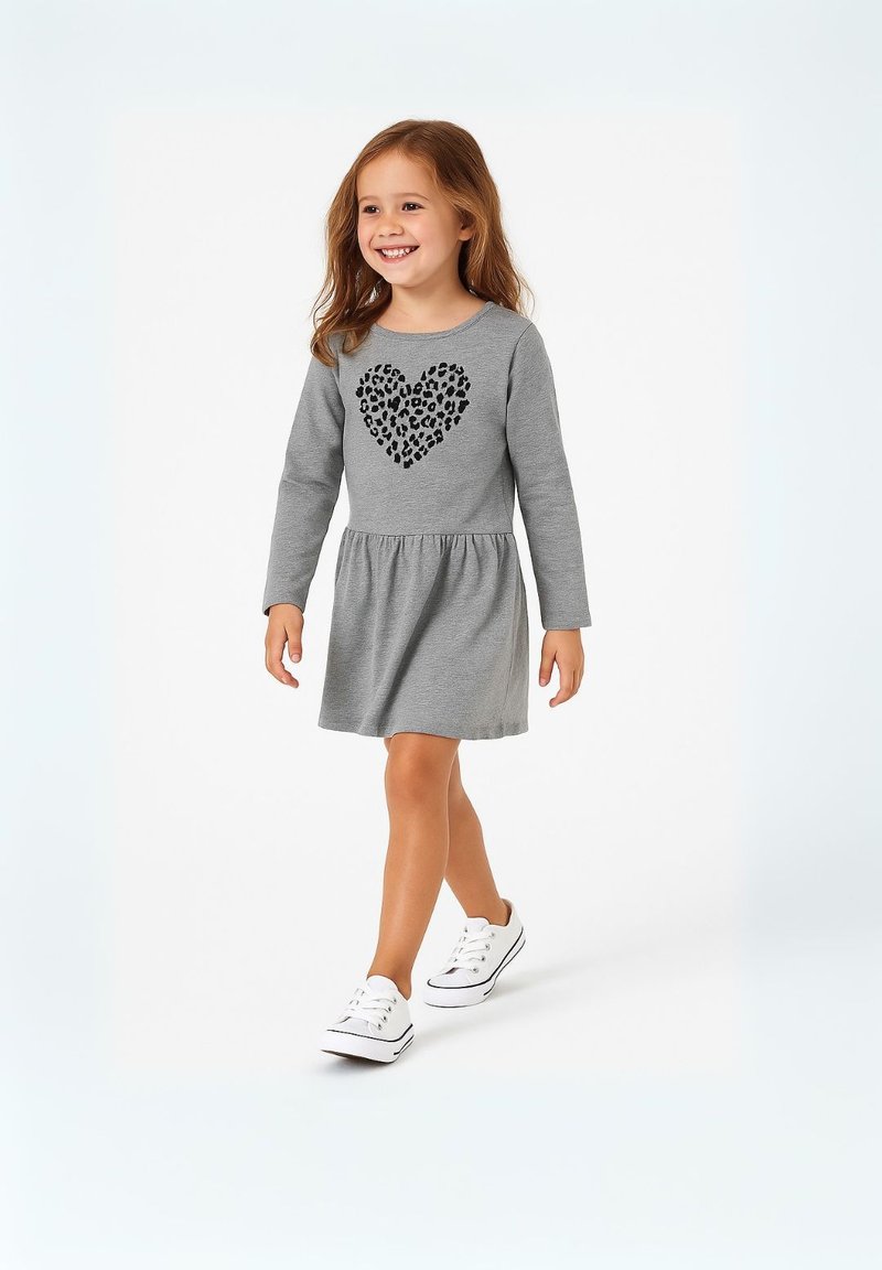 Una giovane ragazza sorridente che cammina, indossando un vestito lungo grigio con un design a cuore leopardato nero e sneakers bianche su uno sfondo bianco.