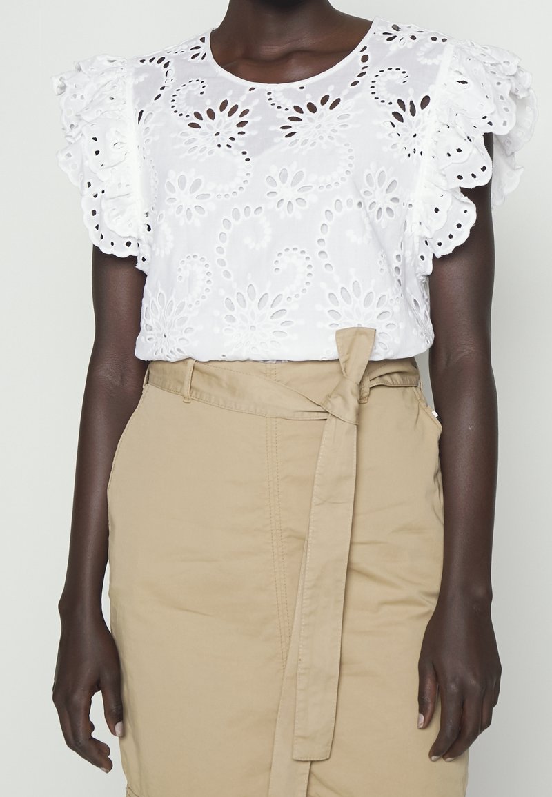 Blouse blanche en dentelle à œillets avec manches courtes froufroutantes, associée à une jupe beige taille haute avec ceinture nouée.