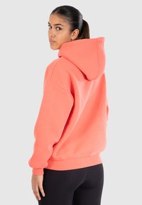 Felpa oversize corallo con cappuccio ampio, polsini e orlo a costine. Texture morbida, design casual, vista da dietro con uno sfondo neutro.