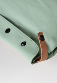 Grünes Canvas-Tasche mit glatter Textur, ausgestattet mit schwarzen Druckknöpfen und einem braunen Lederakzentriemen. Abgerundete Kanten und sichtbare Nähte.