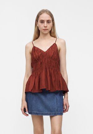 Jeune femme aux longs cheveux blonds porte un chemisier sans manches couleur rouille froncé et une jupe en jean bleue sur un fond blanc uni.