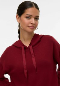 Sudadera burdeos hecha de un tejido suave con capucha con cordones. Presenta una textura lisa y un corte relajado. Diseño sencillo sin patrones ni acentos.