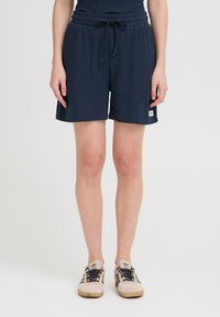 Woman wearing navy ribbed shorts with drawstring and beige sneakers with black stripes, standing against a plain light background.