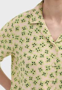 Korte mouwen shirt van een lichtroze stof, met een herhalend bloemmotief in groen en zwart, met een notch-kraag en knoopsluiting.