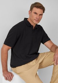 Schwarzes Polohemd mit klassischem Design, kurzen Ärmeln und Kragen. Das Modell kombiniert es mit hellbeigen Hosen. Glatte Textur.