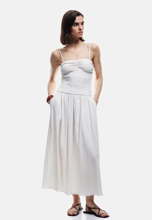 Kobieta w białej sukience maxi bez rękawów z cienkimi ramiączkami, stojąca z rękami w kieszeniach, w czarnych, płaskich sandałach na paski.