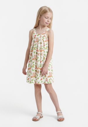 Niña joven con vestido blanco sin mangas con patrones coloridos de frutas y osos de peluche, llevando sandalias blancas, de pie con la mirada hacia un lado.