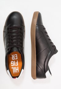 Černé syntetické kožené tenisky s hnědými gumovými podrážkami, které mají šest očíček, jednoduché prošívání a výrazné logo na oranžové stélce.