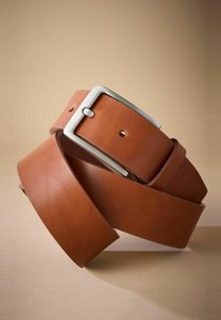 Ceinture en cuir marron avec une boucle en métal argenté, présentant une texture lisse et des trous réglables. Design enroulé sur fond beige.