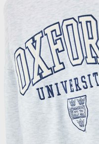 Svijetlosiva tkanina s natpisom "Oxford University" u tamnoplavim blokovima i grbom koji sadrži krunice i tekst o knjigama.