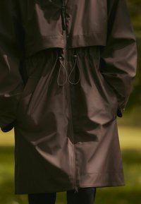 Giacca impermeabile marrone con design corto, cordino elastico in vita e tasche laterali; texture liscia e opaca con chiusura a zip.