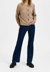 Cardigan beige lavorato a maglia con scollatura rotonda e bottoni neri, abbinato a jeans a palazzo blu scuro e stivali con piattaforma neri. Texture liscia.
