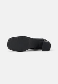 Bota negra de tacón con suela de goma texturizada, que presenta un tacón cuadrado y parte superior lisa. Detallada con suaves ranuras y un acabado mate.