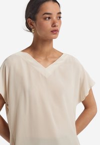 Jasne beżowe bluzka z dekoltem w serek, krótkimi rękawami i luźnym krojem. Miękka, gładka tkanina z delikatnym połyskiem. Brak widocznych wzorów.