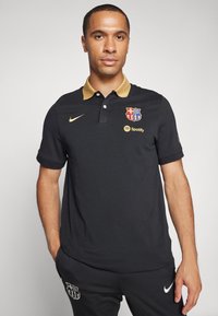 Polo nero con colletto dorato, dotato di logo del Barcellona e simbolo di Spotify. Realizzato in tessuto liscio e traspirante; maniche corte.