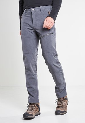 Homme portant un pantalon de plein air gris avec une poche zippée sur la cuisse et des chaussures de randonnée marron, debout devant un fond clair uni.