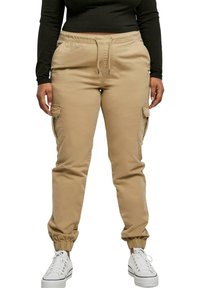Beige cargo pants with elastic waistband and ankle cuffs, featuring side pockets and a smooth, durable fabric. Paired with white sneakers.