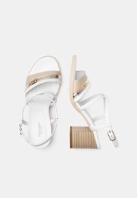 Sandales à talons blanches et beiges avec deux bandes horizontales, une fermeture à boucle et des talons blocs beiges striés. Matériau lisse et brillant avec des accents dorés.