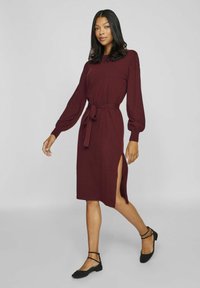 Långärmad klänning i bordeauxfärgad tyg med midjebälte och slits på sidan. Matchas med svarta ballerinas för en komplett look.
