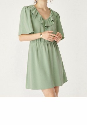 Frau in einem kurzärmligen, hellgrünen Kleid mit Rüschendekolleté und elastischer Taille, posiert mit vor der Brust verschränkten Händen.