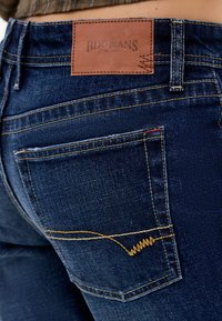 Dark indigo denim jeans with a leather waistband patch, yellow stitching, and a distinctive wavy pattern on the back pocket.