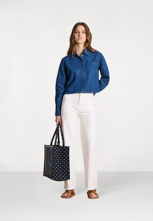 Femme portant une veste en jean bleu, un pantalon blanc, des sandales marron, tenant un sac fourre-tout noir à motifs sur un fond uni.