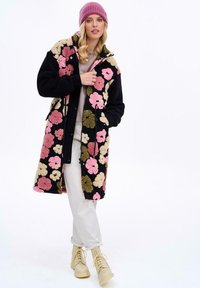 Dlhý kabát s kvetinovým vzorom v čiernej farbe, s pastelovými ružovými a zelenými kvetmi, kapucňou a zapínaním na cvoky. Vyzerajú skvele s béžovými šnurovacími čižmami.