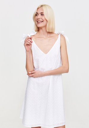 Uśmiechnięta kobieta w białej, bezrękawnikowej sukience z ramiączkami na ramionach stoi na jednolitym tle, delikatnie dotykając swojego ramienia.