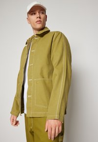 Chaqueta verde oliva de algodón, con cierres a presión, costuras blancas y dos bolsillos frontales. Estilizada con una camiseta blanca debajo.